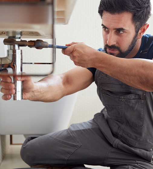 A man repairs a bathroom sink, concentrating on the plumbing while surrounded by tools and fixtures.