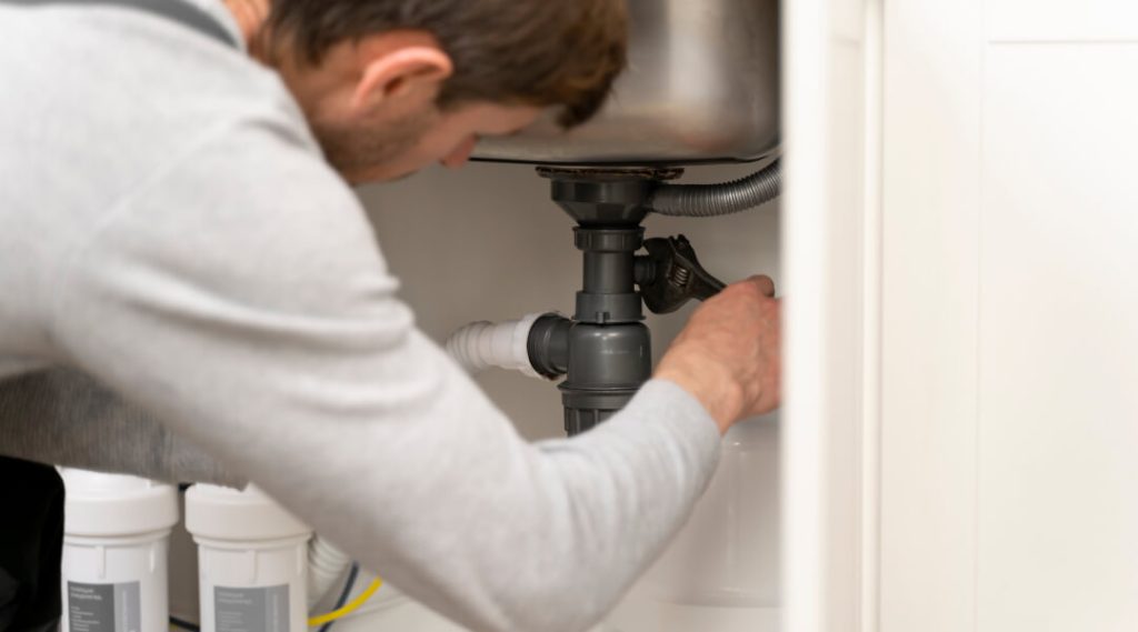 A close-up of a person's hands using a classic black rubber plunger with a wooden handle to effectively clear a blocked drain in a modern stainless steel kitchen sink.