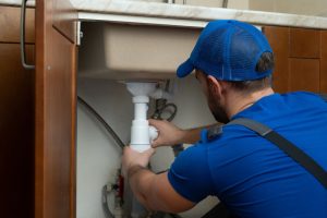 A close-up view of a technician's hands using a wrench to repair and clear a clogged PVC drainage pipe under a modern kitchen sink, illustrating when it's time to call a professional.