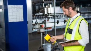 Technicien en gilet de sécurité haute visibilité contrôlant une machine industrielle avec un appareil manuel.